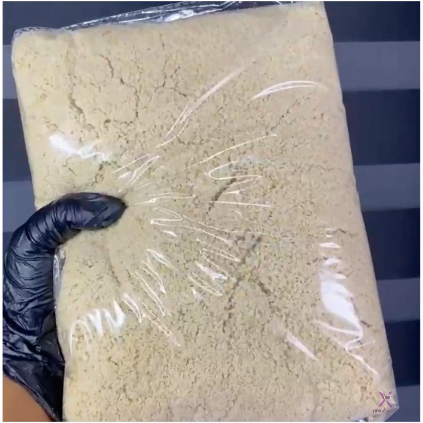 Block of beige material wrapped in clear plastic with a black gloved hand holding it, against a blue and white striped background.